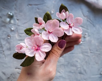 May include: A cluster of delicate pink paper flowers with green leaves, held in a hand. The flowers have a realistic appearance and are perfect for adding a touch of spring to any occasion.