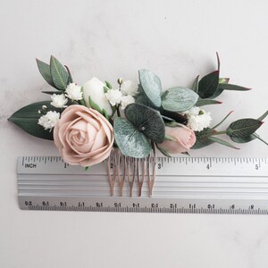 May include: A floral hair comb featuring a blush rose, white flowers, and green leaves. The comb includes a ruler for size reference. The arrangement includes eucalyptus and small white flowers, creating a delicate, natural look.