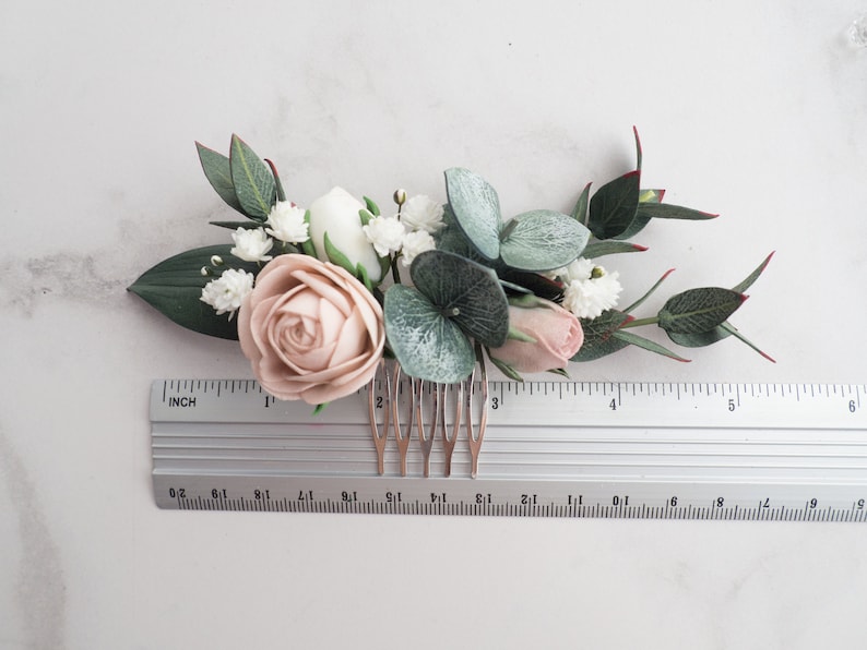 May include: A floral hair comb featuring a blush rose, white flowers, and green leaves. The comb includes a ruler for size reference. The arrangement includes eucalyptus and small white flowers, creating a delicate, natural look.