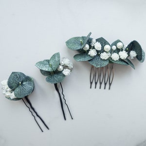 Op de afbeelding: Drie haarclips met groene bladeren en witte bloemen. De grootste clip heeft een kam, de andere twee hebben pinnen.