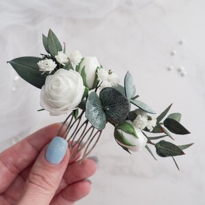 May include: A floral hair comb featuring white roses, buds, and small white flowers, accented with green leaves and eucalyptus. The comb is made of gold-colored metal. A delicate accessory for weddings or special occasions.