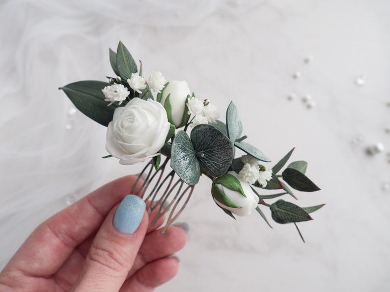 May include: A floral hair comb featuring white roses, buds, and small white flowers, accented with green leaves and eucalyptus. The comb is made of gold-colored metal. A delicate accessory for weddings or special occasions.