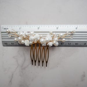 May include: A gold-toned hair comb featuring white floral accents, round white pearls, and clear crystal beads on thin wire branches. A metal ruler placed behind the comb shows a total width of approximately 4 inches for the decorative top section.