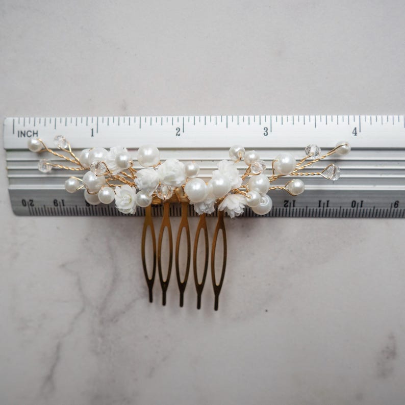 May include: A gold-toned hair comb featuring white floral accents, round white pearls, and clear crystal beads on thin wire branches. A metal ruler placed behind the comb shows a total width of approximately 4 inches for the decorative top section.