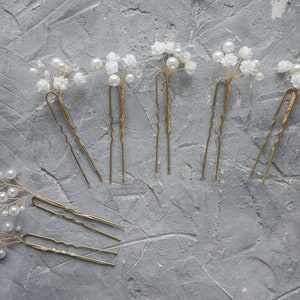May include: Seven gold hair clips with white pearl and flower embellishments. The clips are arranged in a semi-circle on a gray textured surface.