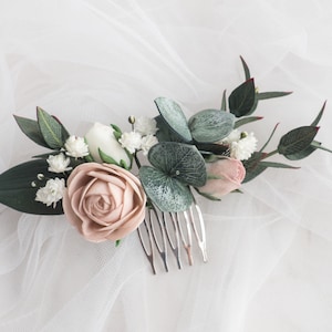 May include: A floral hair comb featuring a large blush rose, white and pink buds, and green leaves. The comb is made of silver metal and is designed for bridal or special occasion hairstyles.