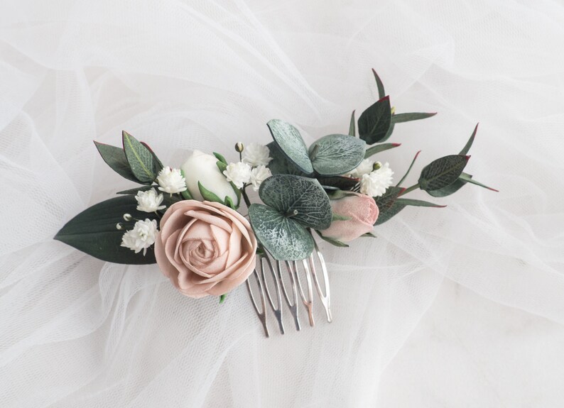 May include: A floral hair comb featuring a large blush rose, white and pink buds, and green leaves. The comb is made of silver metal and is designed for bridal or special occasion hairstyles.