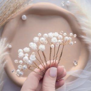 May include: A set of gold-toned hair pins featuring white floral accents, round white faux pearls, and small clear crystal beads. The decorative elements are attached to thin, flexible gold-colored wire stems that extend from the U-shaped hair pins.