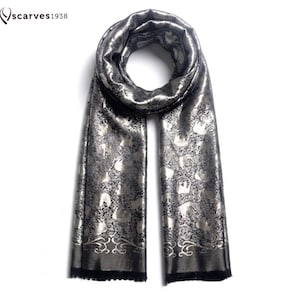 May include: A metallic silver and black scarf featuring a repeating pattern of elephants and intricate floral scrollwork. The scarf includes the text "scarves1938" in the top left corner, has a draped loop at the top, and ends with a fringe.