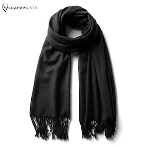 Puede incluir: Una bufanda negra sólida con una textura suave y lisa, que presenta flecos largos y finos en ambos extremos. En la esquina superior izquierda aparece el texto "scarves1938" junto a un icono de logotipo estilizado. La bufanda está doblada.