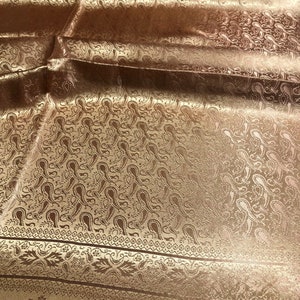 May include: A shimmering bronze-colored fabric featuring an intricate woven paisley pattern throughout. The textile displays a distinct horizontal border with floral motifs and geometric lines, showcasing a reflective, satin-like finish.