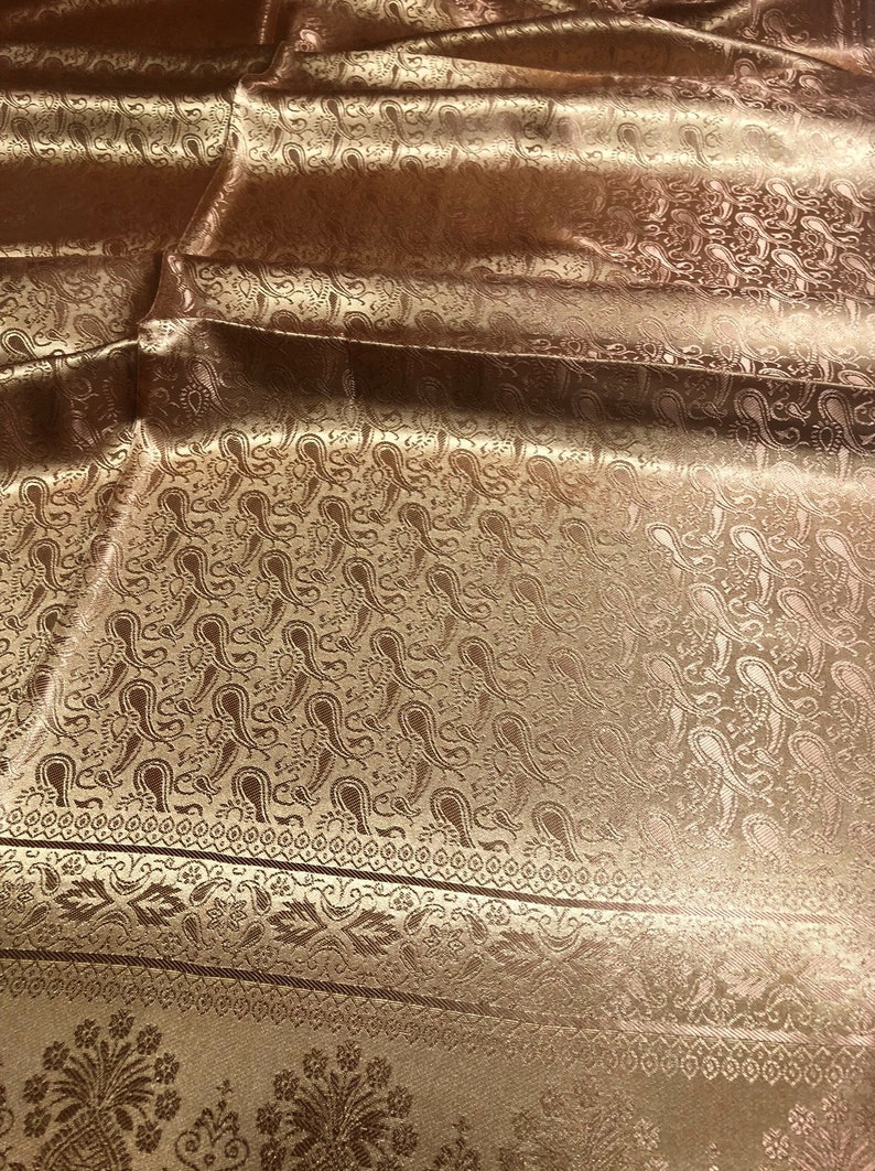 May include: A shimmering bronze-colored fabric featuring an intricate woven paisley pattern throughout. The textile displays a distinct horizontal border with floral motifs and geometric lines, showcasing a reflective, satin-like finish.