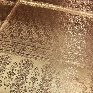 May include: A bronze-colored fabric featuring a woven paisley pattern and a decorative border with floral motifs. The material has a subtle sheen and includes a black rectangular tag in the corner with the white text 100% SILK and care symbols.
