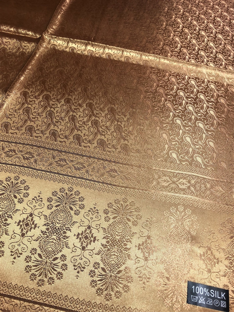 May include: A bronze-colored fabric featuring a woven paisley pattern and a decorative border with floral motifs. The material has a subtle sheen and includes a black rectangular tag in the corner with the white text 100% SILK and care symbols.