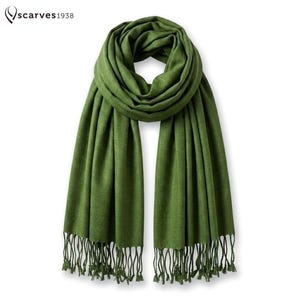 Peut inclure: Une écharpe vert olive unie présentant une texture tissée douce et des franges le long des bords inférieurs. L'écharpe est drapée en boucle sur le dessus avec deux longues extrémités pendantes. Le texte "scarves1938" est imprimé en haut à gauche.