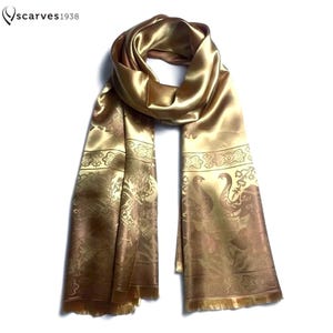 Puede incluir: Una bufanda de satén de color dorado con un patrón repetitivo de elefantes y bordes florales decorativos. La bufanda incluye el texto "scarves1938" en la esquina superior izquierda y tiene un acabado liso y reflectante con flecos.