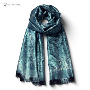 Puede incluir: Una bufanda de color turquesa con un estampado de cachemira y bordes con flecos. La bufanda tiene una textura brillante y sedosa y está drapeada para mostrar su diseño. La marca "scarves1938" está en la esquina superior izquierda.