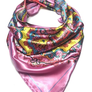 May include: A square satin scarf featuring a vibrant paisley pattern in shades of pink, yellow, and turquoise. The scarf is folded into a triangular shape, displaying a smooth, reflective surface with intricate decorative motifs and a thin blue border.