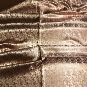 May include: A shimmering bronze-colored fabric featuring a repeating paisley pattern woven into the material. The textile has a smooth, reflective surface with deep folds and creases that create varying light and shadow across the intricate design.