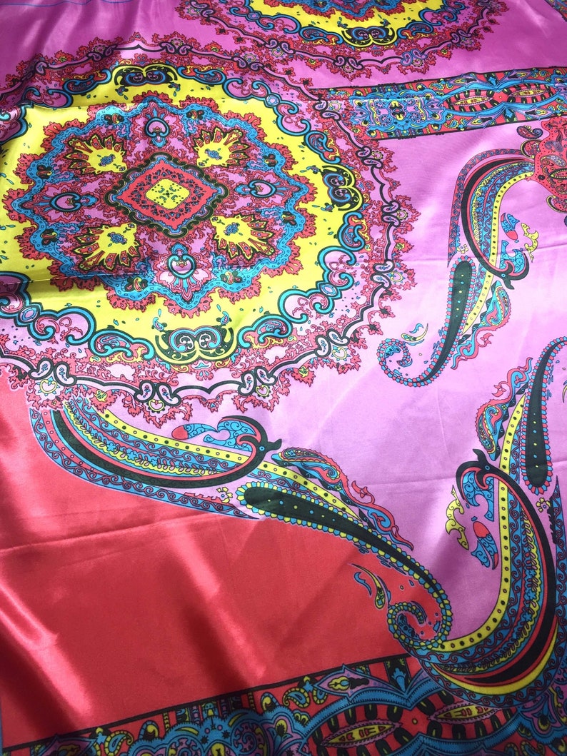 May include: A vibrant fabric featuring a large, intricate circular mandala pattern in yellow, pink, and blue. The design includes detailed paisley motifs and geometric shapes, set against a smooth, shiny, satin-like material with a bright pink base.