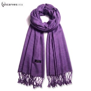May include: A solid purple scarf with a soft, woven texture and fringed ends. The scarf features a small black rectangular label stitched onto the fabric. The text "scarves1938" is visible in the upper left corner of the image area.