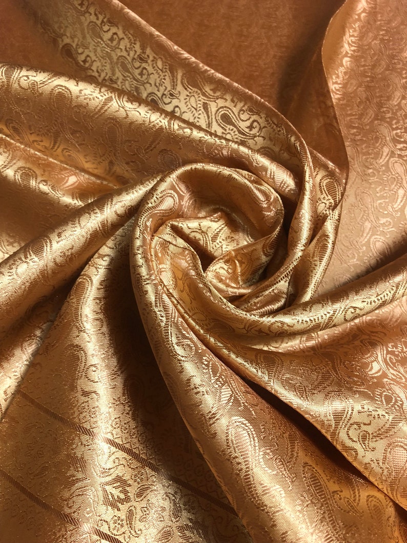 May include: A shimmering copper-colored fabric featuring a woven paisley pattern. The material is gathered into a central swirl, highlighting the reflective quality of the textile and the intricate, repeating teardrop-shaped motifs across the surface.