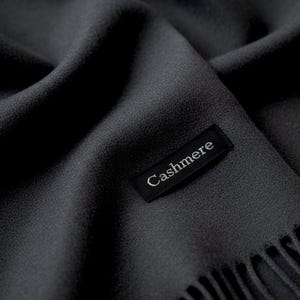 Puede incluir: Una bufanda de color gris carb&oacute;n oscuro con una superficie de textura fina y suave. Una etiqueta rectangular de tela negra est&aacute; cosida sobre la tela, con la palabra Cashmere bordada en hilo blanco. El borde de la bufanda tiene flecos.