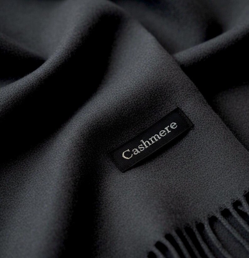 Puede incluir: Una bufanda de color gris carb&oacute;n oscuro con una superficie de textura fina y suave. Una etiqueta rectangular de tela negra est&aacute; cosida sobre la tela, con la palabra Cashmere bordada en hilo blanco. El borde de la bufanda tiene flecos.