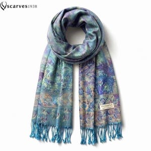 Peut inclure: Une écharpe rectangulaire présentant un motif cachemire et floral multicolore dans des tons de bleu, violet et vert. L'écharpe a des extrémités à franges et une petite étiquette blanche indiquant "PASHMINA 100% Cashmere". Le logo "scarves1938" est visible.