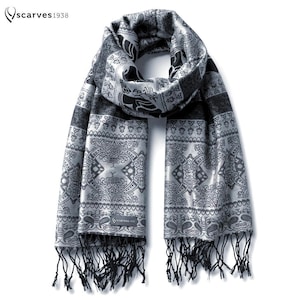 Op de afbeelding: Een zilveren en zwarte sjaal met een herhalend ingewikkeld patroon van geometrische vormen en gestileerde motieven. De sjaal heeft een rechthoekige vorm met een klein rechthoekig label met de tekst scarves1938 en zwarte franjes aan de onderkant.
