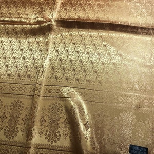 May include: A gold-colored silk fabric featuring a repeating paisley pattern on the upper section and a decorative floral border on the lower section. A small black rectangular tag in the corner displays the text 100% SILK with care icons.