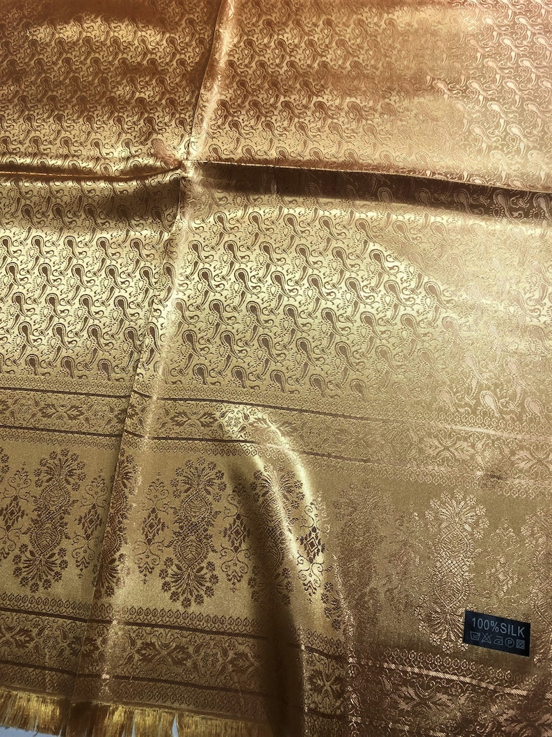May include: A gold-colored silk fabric featuring a repeating paisley pattern on the upper section and a decorative floral border on the lower section. A small black rectangular tag in the corner displays the text 100% SILK with care icons.