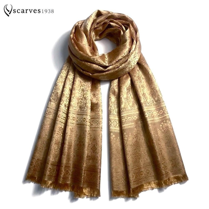 May include: A gold-toned scarf featuring an intricate woven pattern with floral and geometric motifs. The scarf has a soft sheen, a draped neckline, and fringed edges at the bottom. The text "scarves1938" is visible in the upper left corner.