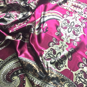 May include: A pink satin fabric with a paisley pattern in black, gold, and white. The fabric is folded and draped, showing the intricate details of the design.