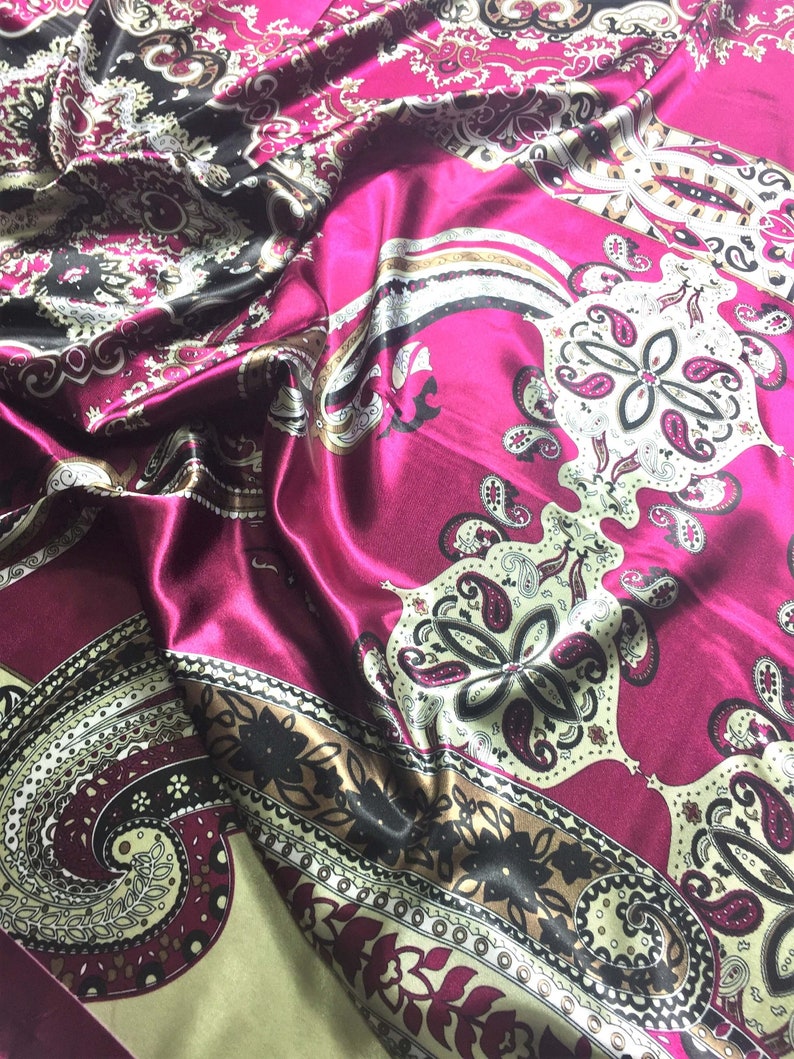 May include: A pink satin fabric with a paisley pattern in black, gold, and white. The fabric is folded and draped, showing the intricate details of the design.