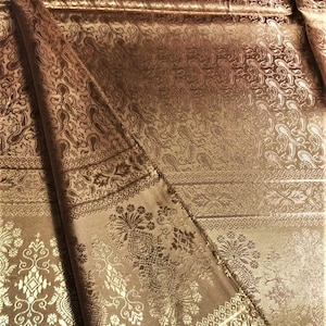 May include: A metallic bronze fabric featuring an intricate woven jacquard pattern. The textile displays repeating paisley motifs, ornate floral borders, and geometric decorative bands with a subtle sheen that highlights the detailed texture.