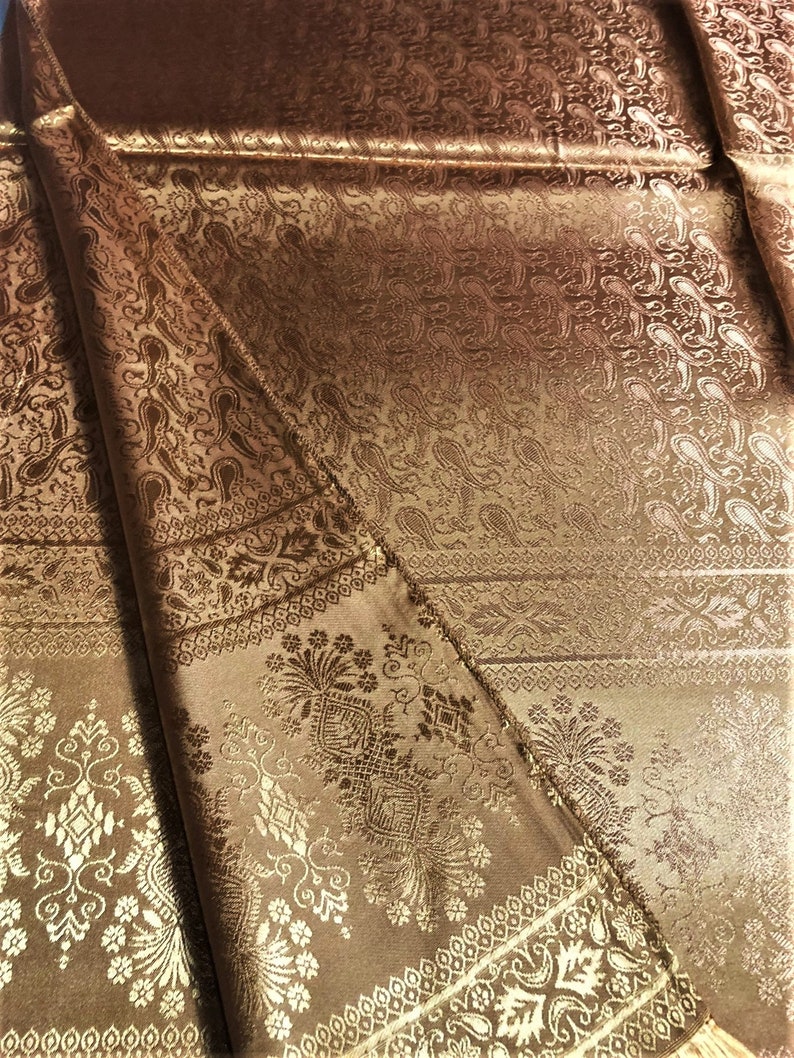 May include: A metallic bronze fabric featuring an intricate woven jacquard pattern. The textile displays repeating paisley motifs, ornate floral borders, and geometric decorative bands with a subtle sheen that highlights the detailed texture.