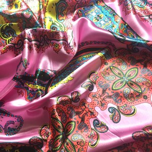 May include: A satin scarf featuring a vibrant paisley pattern in shades of pink, yellow, turquoise, and coral. The fabric has a smooth, reflective surface with intricate floral and teardrop motifs arranged in a repeating, colorful design.