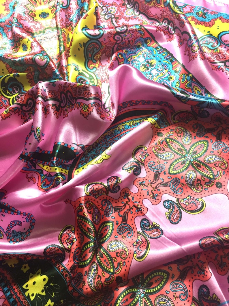 May include: A satin scarf featuring a vibrant paisley pattern in shades of pink, yellow, turquoise, and coral. The fabric has a smooth, reflective surface with intricate floral and teardrop motifs arranged in a repeating, colorful design.