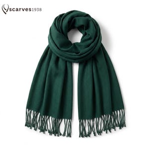 Puede incluir: Una bufanda de color verde bosque oscuro sólido con una textura suave y lisa y un diseño en bucle. La bufanda tiene flecos largos y finos en ambos bordes inferiores. El texto "scarves1938" está impreso en negro en la esquina superior izquierda.