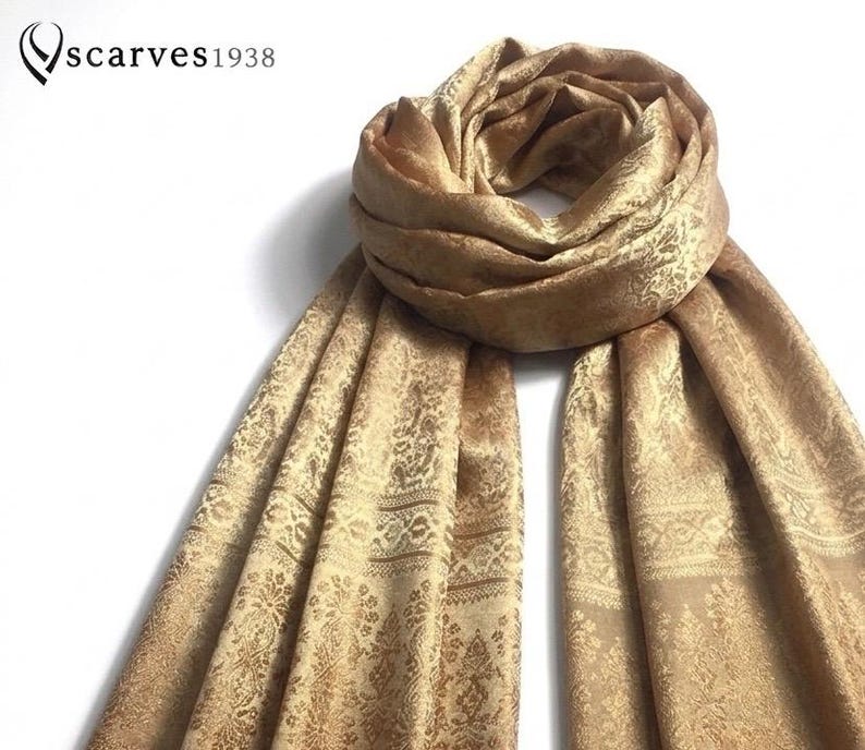May include: A gold-colored scarf featuring an intricate, repeating jacquard pattern with floral and geometric motifs. The scarf is draped in a circular loop at the top with two long ends hanging down. The text "scarves1938" appears in the top left corner.
