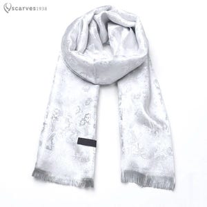 Pode incluir: Um cachecol branco com um padrão paisley jacquard tonal e um brilho subtil. O cachecol tem uma forma retangular com extremidades franjadas e uma pequena etiqueta retangular preta presa ao tecido. O texto "scarves1938" aparece no canto superior.