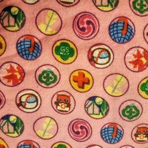 May include: Pink fabric with a repeating pattern of colorful circular patches. The patches feature various designs, including a cross, a globe, a leaf, and the text "GS WAY". The patches are outlined in a darker pink.