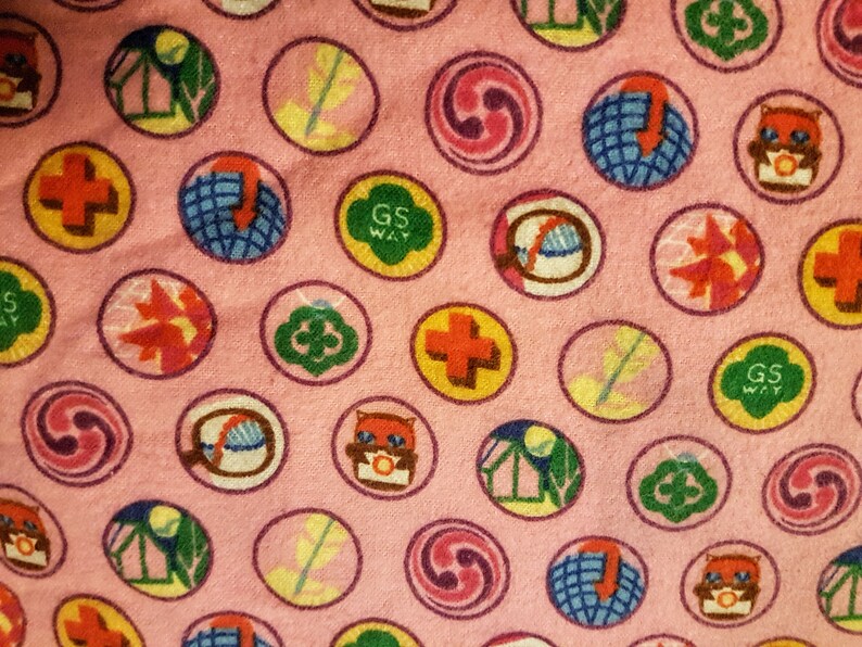 May include: Pink fabric with a repeating pattern of colorful circular patches. The patches feature various designs, including a cross, a globe, a leaf, and the text "GS WAY". The patches are outlined in a darker pink.