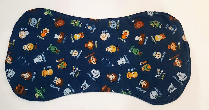 May include: A navy blue baby bib featuring a repeating pattern of Star Wars characters and names. The bib has a unique hourglass shape and is made of soft fabric. Characters include Yoda, C-3PO, and Princess Leia.