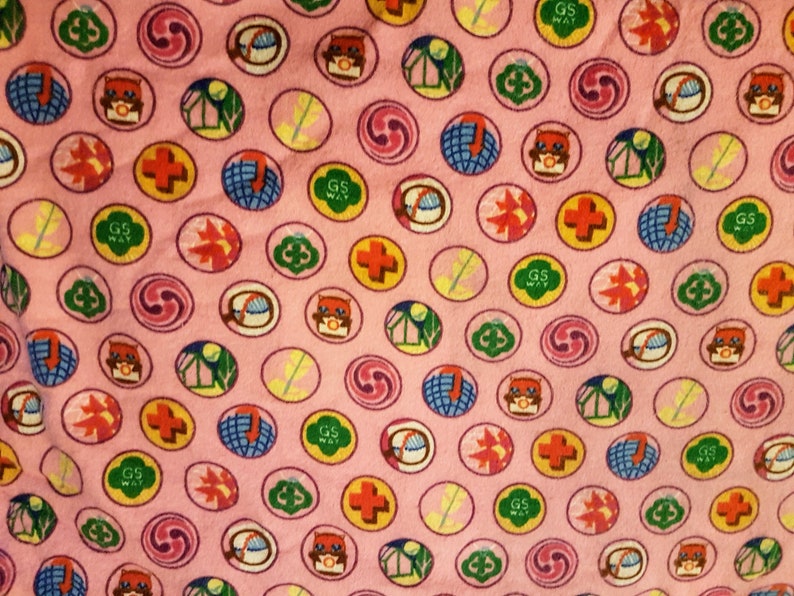 May include: Pink fabric with a repeating pattern of circular designs. Each circle contains various colorful illustrations, including a cross, a swirl, a globe, a leaf, and an owl. The text "GS WAY" is also present.