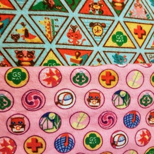 May include: Close-up of two patterned fabrics. The top fabric features triangular shapes with animal illustrations and the text "GS WAY". The bottom fabric is pink with circular designs, including the text "GS WAY" and various symbols.