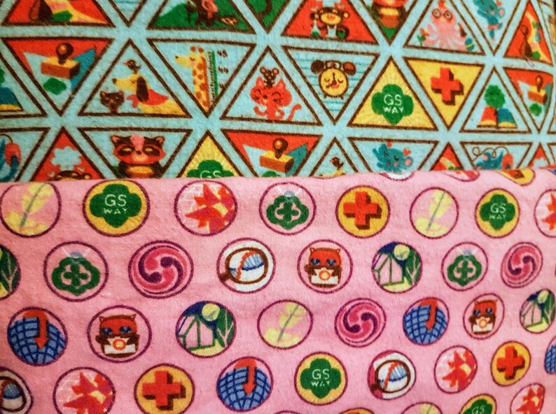 May include: Close-up of two patterned fabrics. The top fabric features triangular shapes with animal illustrations and the text "GS WAY". The bottom fabric is pink with circular designs, including the text "GS WAY" and various symbols.