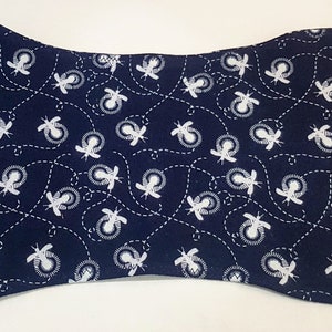May include: Navy blue baby bib with a white bee pattern. The bib is a unique shape, with a curved top and bottom, and a narrower center. The bee design is repeated across the surface.