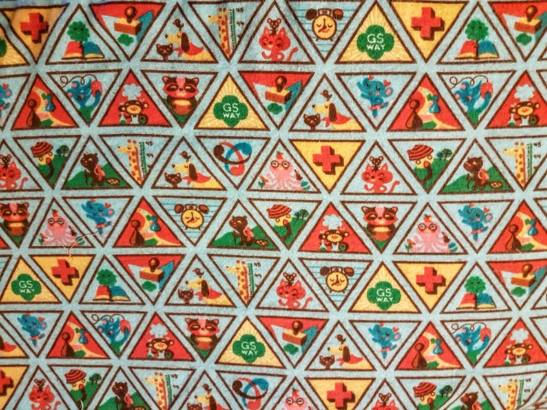 May include: A light blue fabric with a repeating pattern of colorful triangular patches. Each triangle features a different illustration of animals, objects, and the text "GS WAY". The patches are outlined in brown.